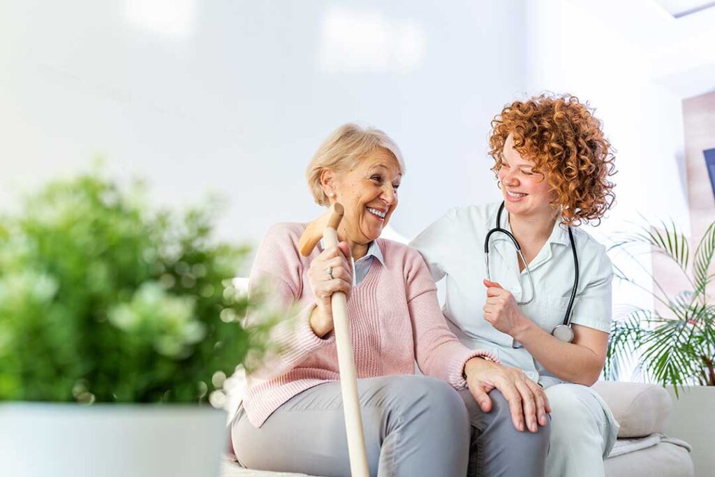 Two seniors chatting and smiling at a table in a sunny, communal living room.